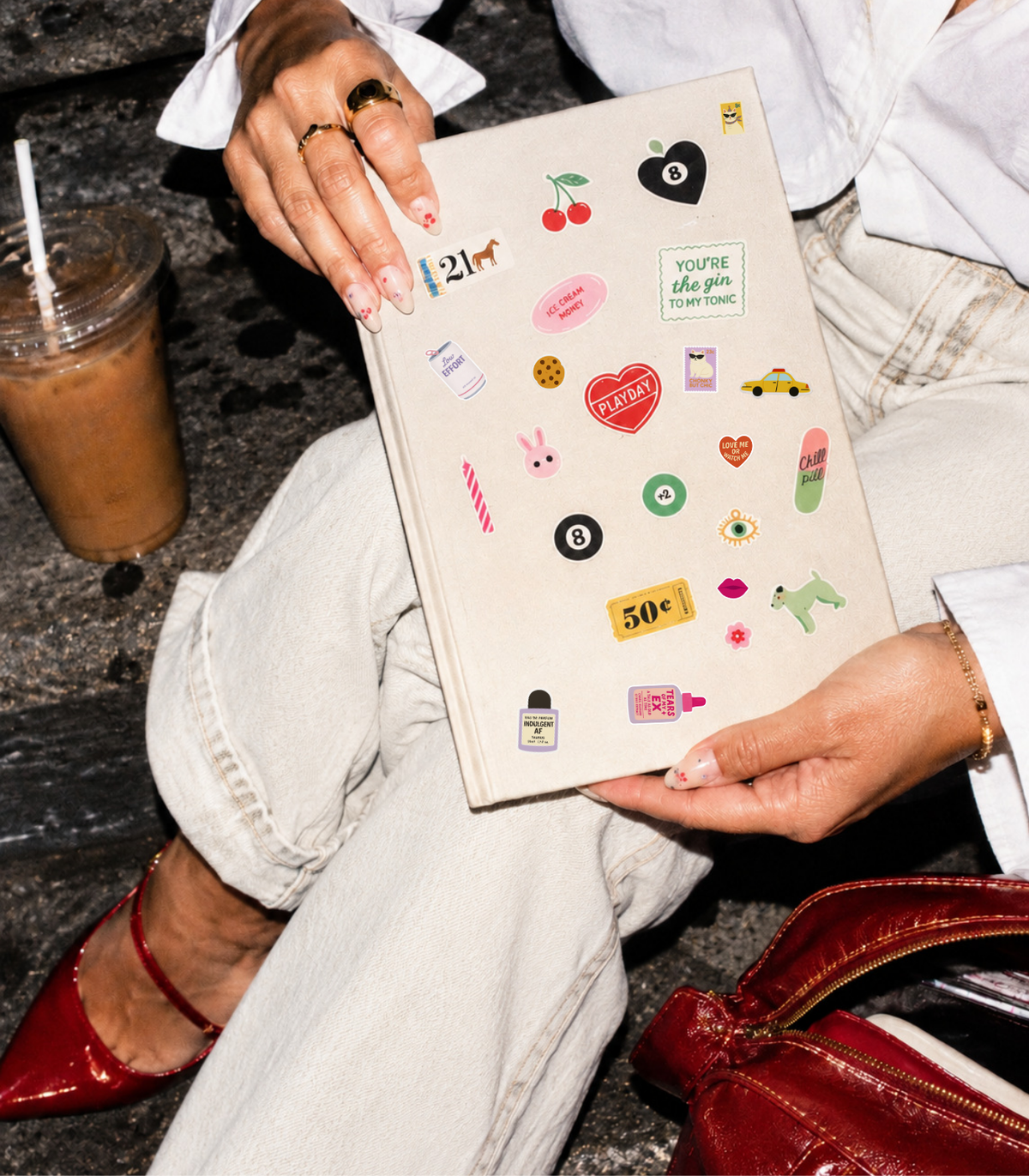 A person in a white shirt and light jeans holds a cream-colored notebook or journal decorated with colorful stickers, seated outdoors next to an iced coffee and a red handbag.