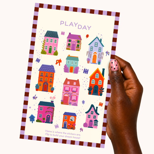 A hand with pink polka-dot nail polish holds a children's activity sticker sheet titled 'PLAYDAY'. The sheet features colorful cartoon houses, small trees, and decorative stickers on an off-white background with a purple and brown border, set against a plain light beige background.