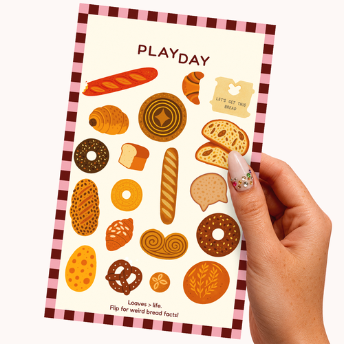 A hand with decorated nails holds a sticker sheet featuring illustrated bread and pastry shapes and the word PLAYDAY at the top, against a plain light background.