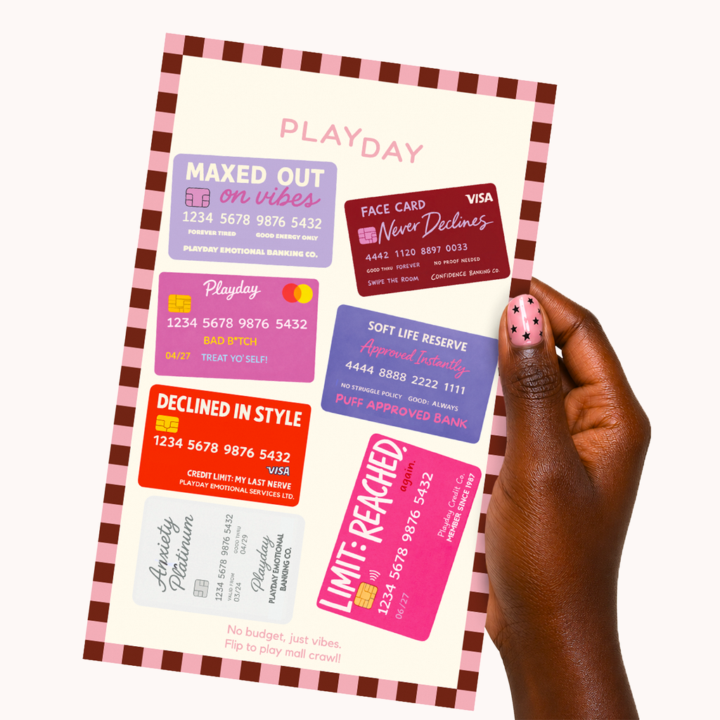 A hand with pink nail polish holds a Playday credit card sticker sheet against a light cream background. The mat features a pink and white checkered border and displays several colorful sample credit cards with humorous finance-themed text like 'MAXED OUT on vibes', 'FACE CARD', 'DECLINED IN STYLE', and 'LIMIT-REACHED'.