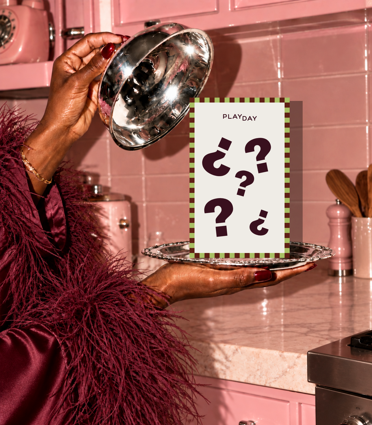 A person in a pink kitchen holds a silver metal bowl over a silver tray, with a card reading 'PLAYDAY' and question marks in the center.