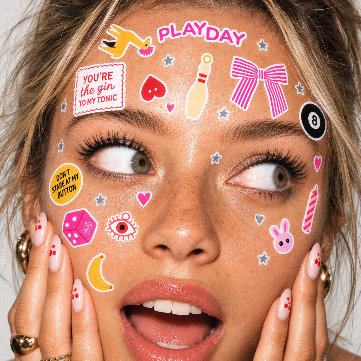 A close-up of a woman's face decorated with colorful playful stickers including the words 'PLAY DAY', 'YOU'RE THE GIR TO MY TONIC', hearts, stars, a bowling pin, a pink bow, a black 8-ball, a rabbit face, and other fun graphics.