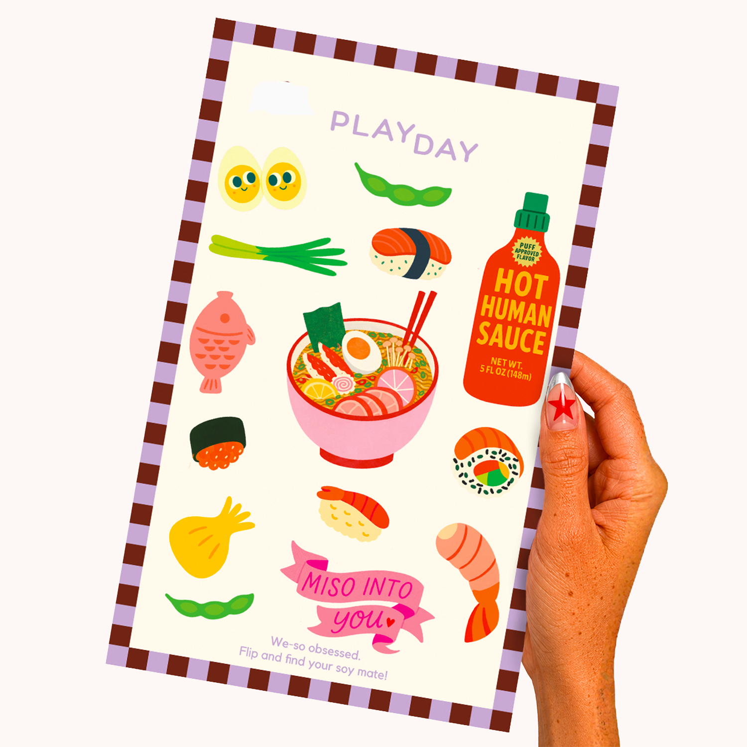 A hand holds a playday sticker sheet featuring colorful Japanese-themed stickers like sushi, ramen, edamame, and a hot sauce bottle on a cream cover with a brown and lavender check border.